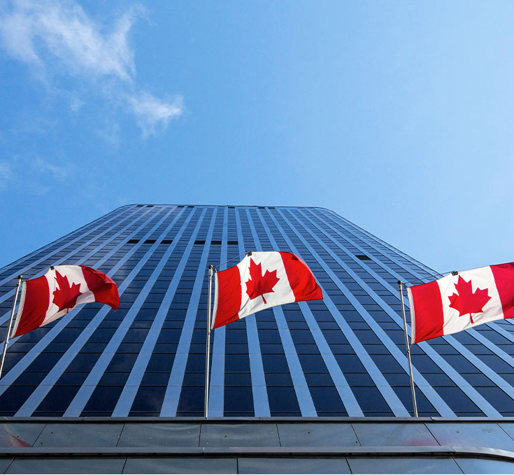 Trois drapeaux canadiens devant une grande tour de bureaux vus d’en bas.