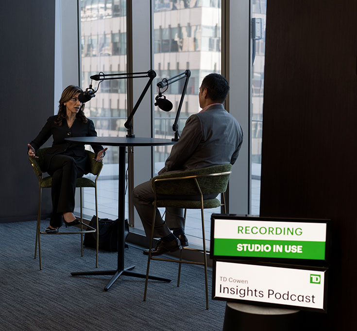 Two business people sitting at a table in an office in front of large podcast microphones. A sign up front says Recording Studio In Use.