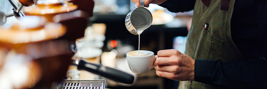 Behind the counter of a coffee shop, a barista pours milk froth into a cup to make a café latte.