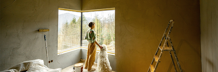 A woman in a home surrounded by supplies for painting a room is petting a dog and looking outside through a window.