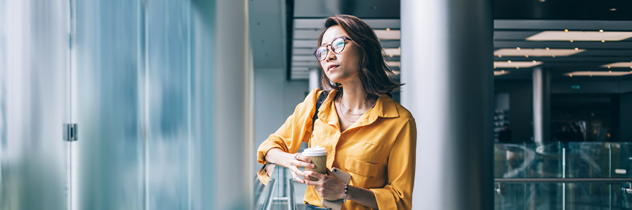 A woman of Asian descent holding a phone and coffee looking out the window.