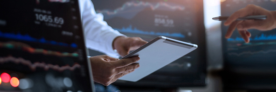 Two people analyzing chart data on a tablet computer and computer monitors.