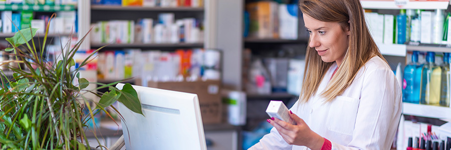 A pharmacist holding a box of drugs and looking up information on a desktop computer in a pharmacy.