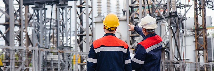 Two workers in hard hats at a power transfer station surrounded by high-power transformers.