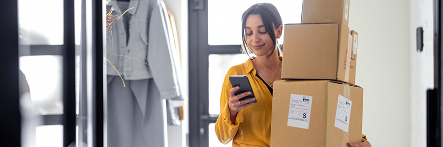 A woman carrying multiple delivery boxes at her front door and looking at her cell phone.