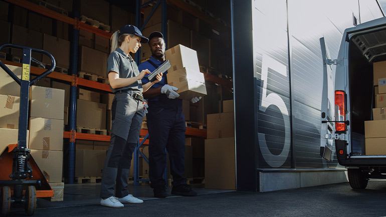 Two parcel delivery people standing in the doorway of a warehouse. One is holding boxes and the other is looking up information on a tablet computer.