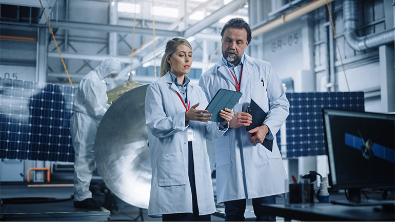 Two engineers in white coats looking over data on a tablet computer in a tech lab.