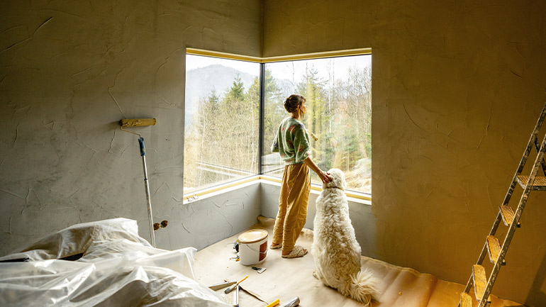 A woman in a home surrounded by supplies for painting a room is petting a dog and looking outside through a window.