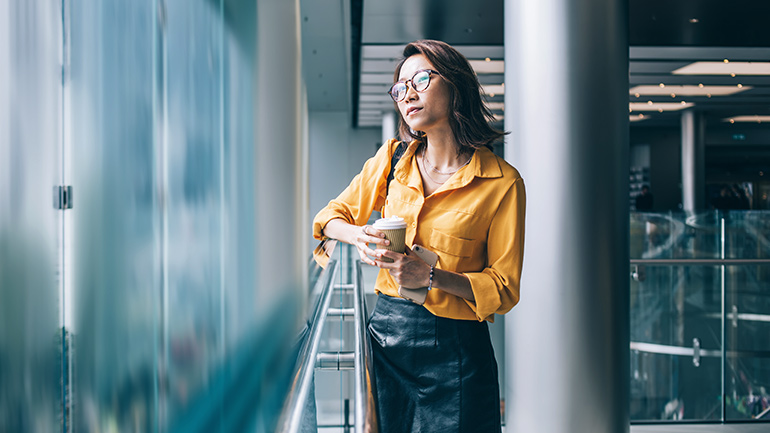 A woman of Asian descent holding a phone and coffee looking out the window.