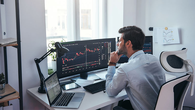 A man sitting at a desk with multiple computer monitors displaying data on large charts.