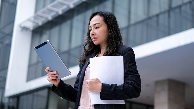 A businessperson holding printouts and looking at information on a tablet computer.